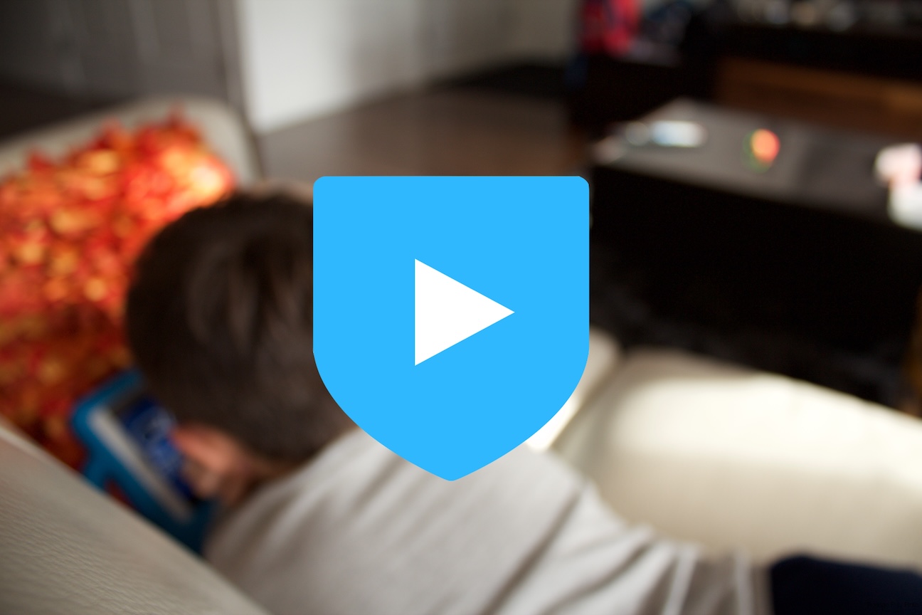 Blue shield with white triangle in the middle. The triangle is tilted to the right, giving the idea that the shield is a play button. The background is a child playing with an iPad on a sofa.
